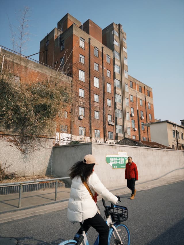 A person rides a bicycle on a street near a large brick building, while another person walks nearby. The sky is clear and blue