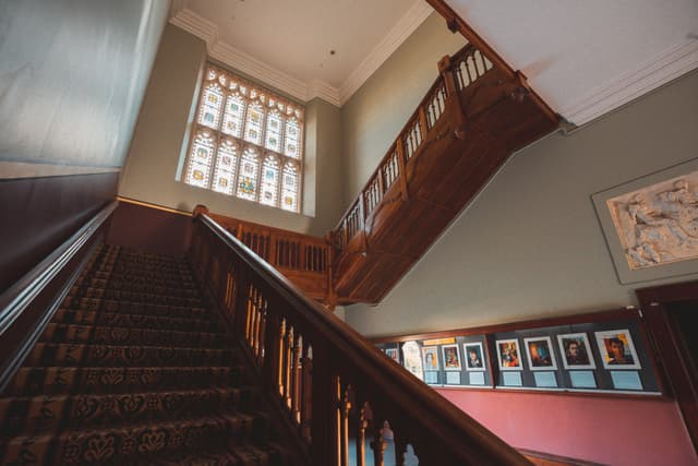 A grand wooden staircase with ornate railings, leading to a landing with large, decorative windows. The walls are adorned with framed pictures and a map