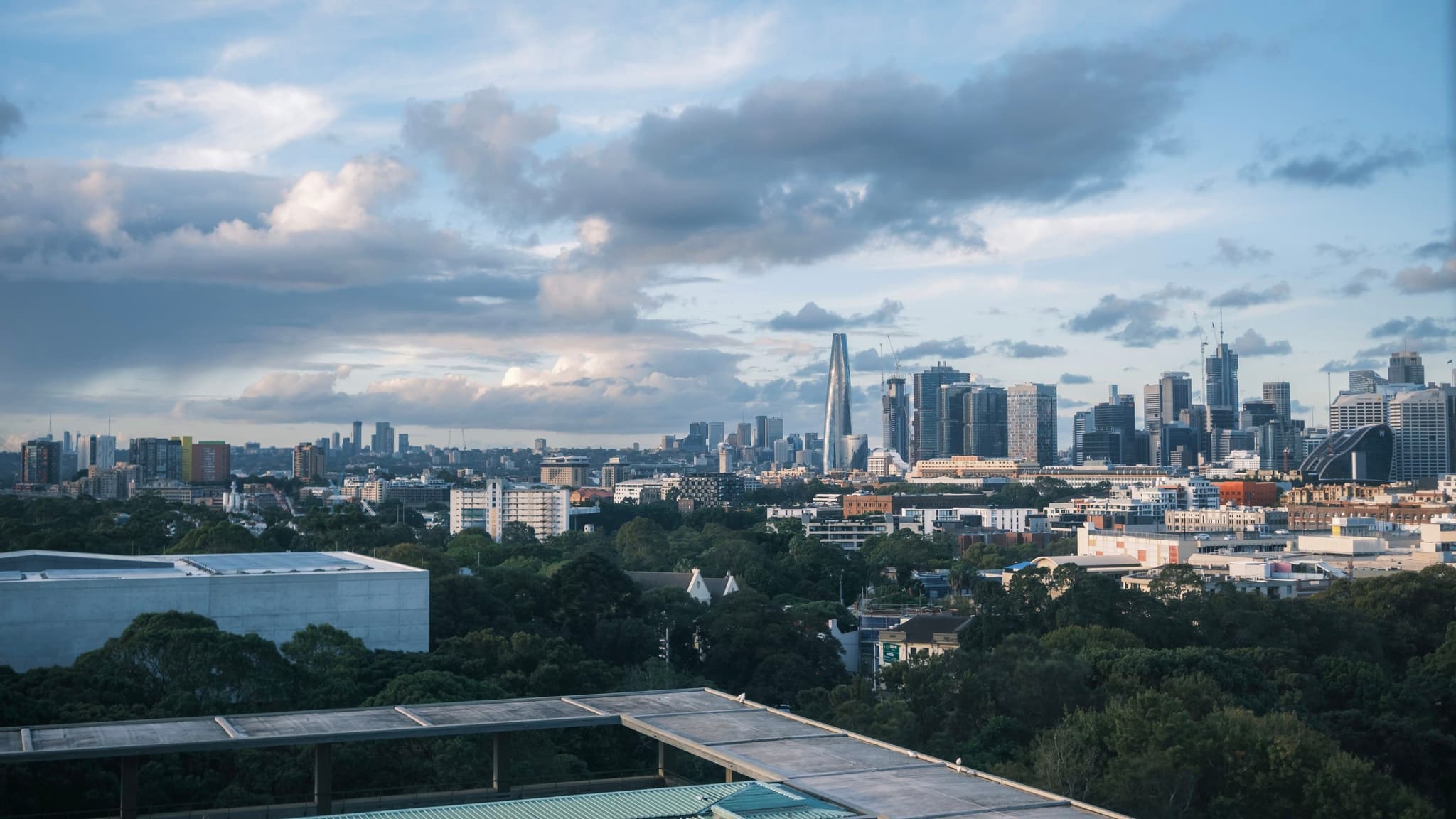 A city skyline with modern skyscrapers under a partly cloudy sky, surrounded by greenery and urban buildings