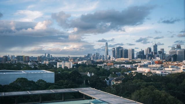A city skyline with modern skyscrapers under a partly cloudy sky, surrounded by greenery and urban buildings