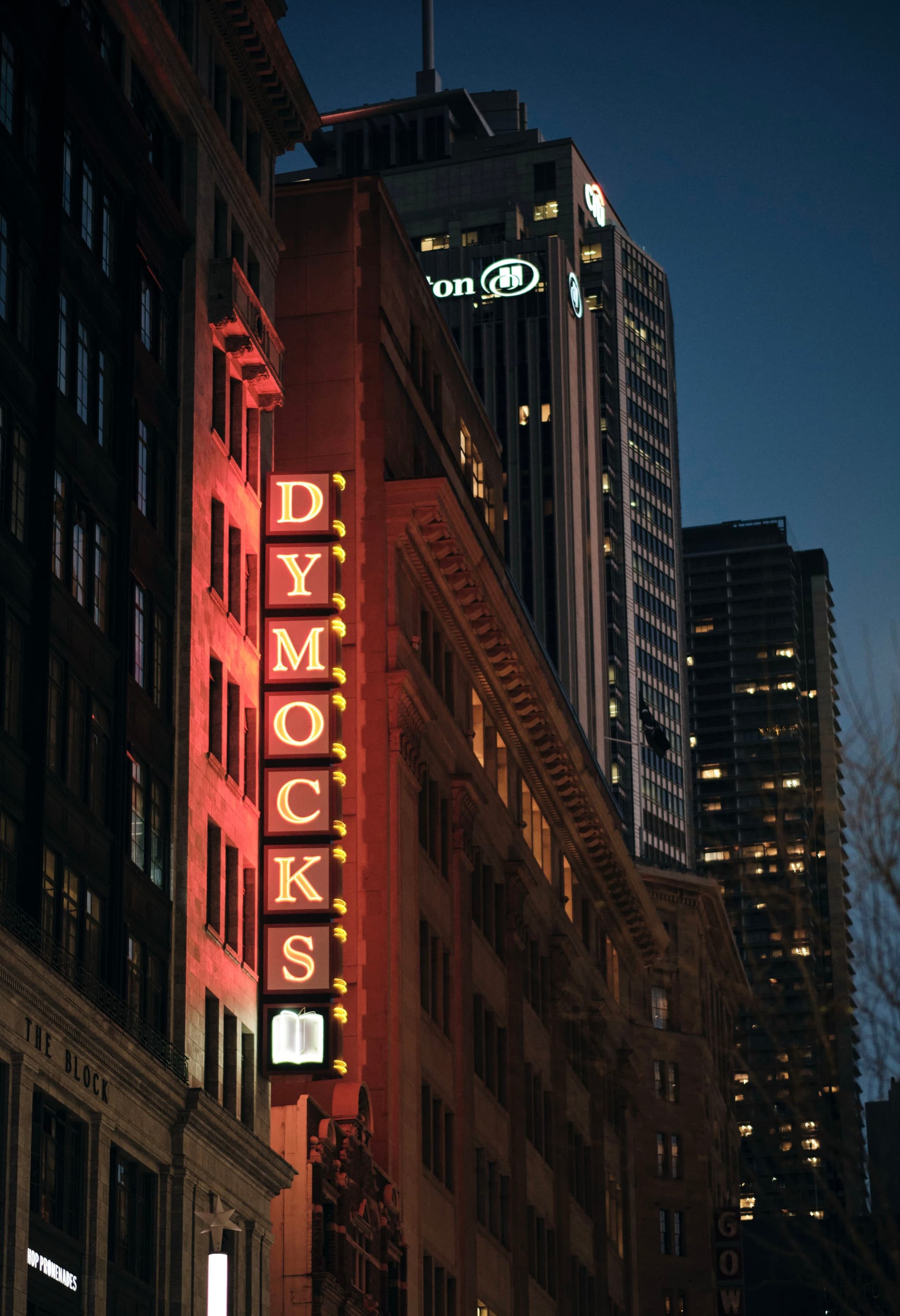A city street at dusk with a prominent vertical Dymocks sign on a building, surrounded by other tall buildings