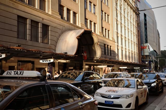 A busy city street with several taxis lined up in front of a large building, featuring a prominent awning and bustling pedestrian activity