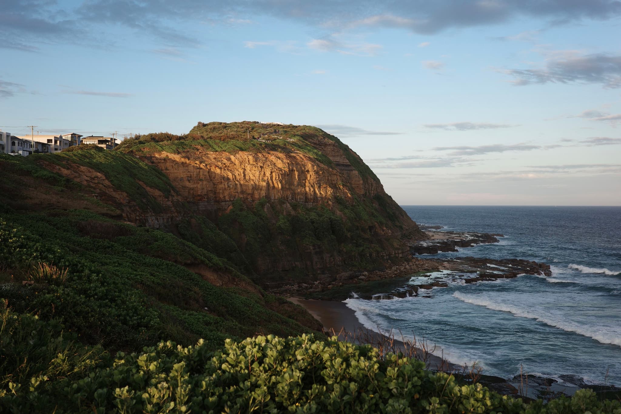 A coastal landscape with a rocky cliff, green vegetation, and waves crashing against the shore under a partly cloudy sky