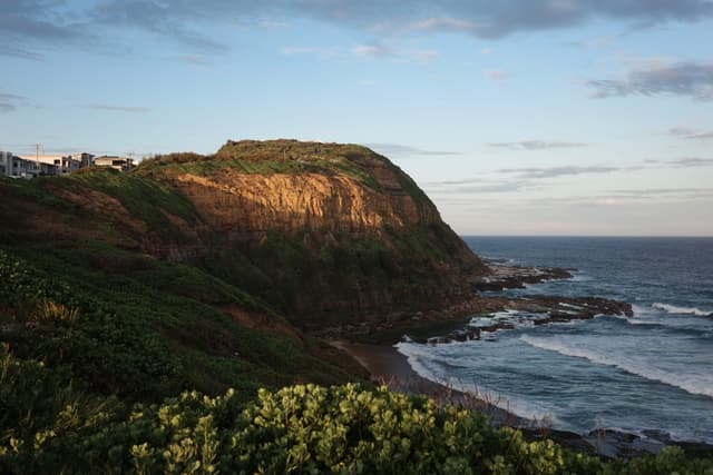 A coastal landscape with a rocky cliff, green vegetation, and waves crashing against the shore under a partly cloudy sky