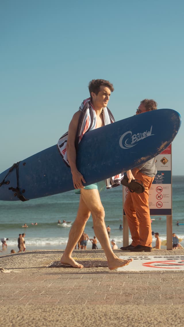 A person carrying a blue surfboard walks along a beach, with the ocean and other beachgoers in the background
