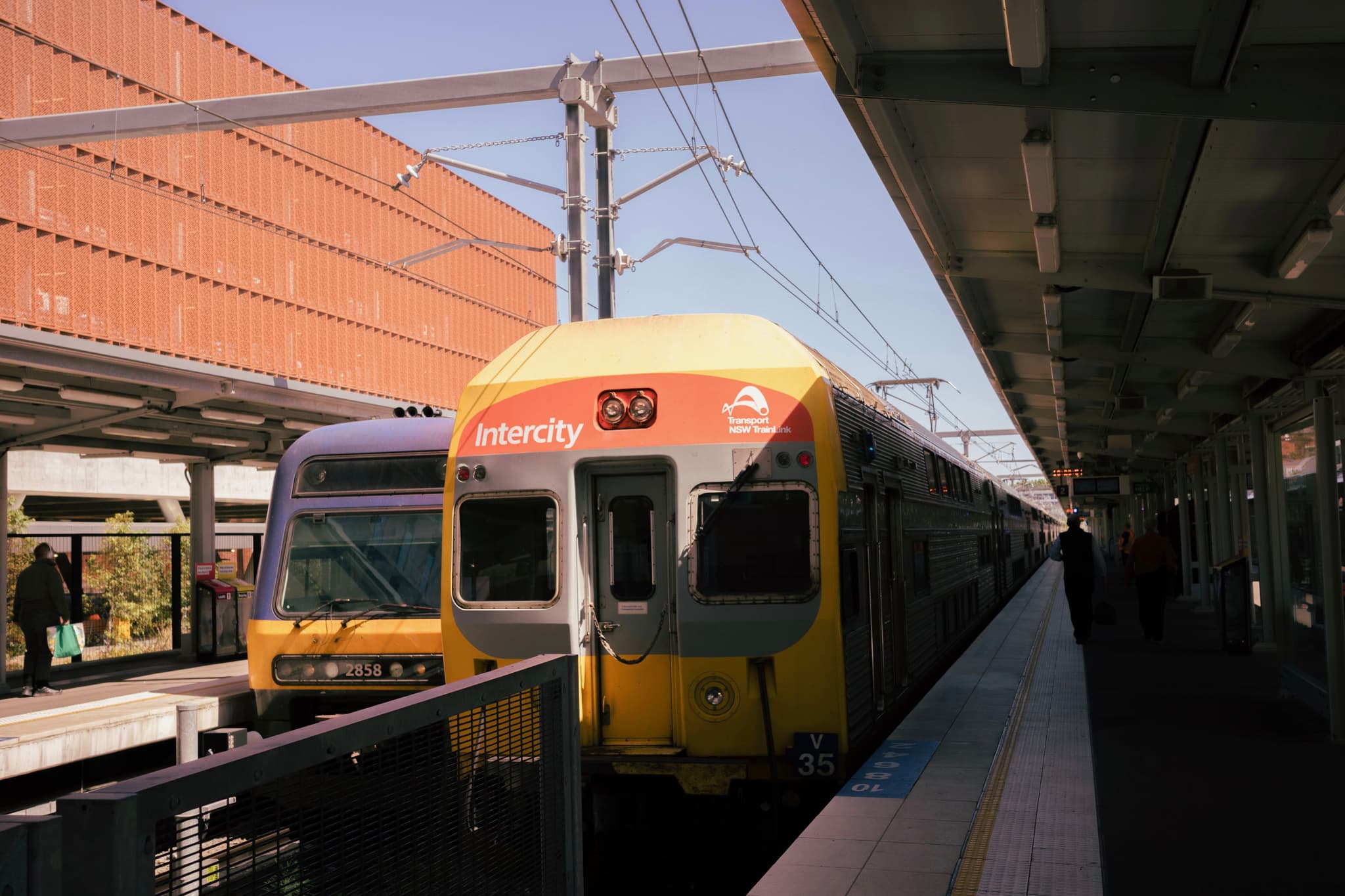 Two trains at a station platform, with one train labeled Lidcombe and overhead power lines visible