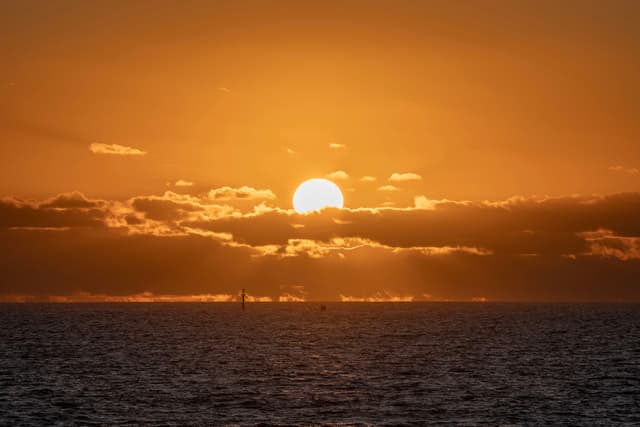 A sunset over the ocean with the sun partially obscured by clouds, casting an orange glow across the sky and water