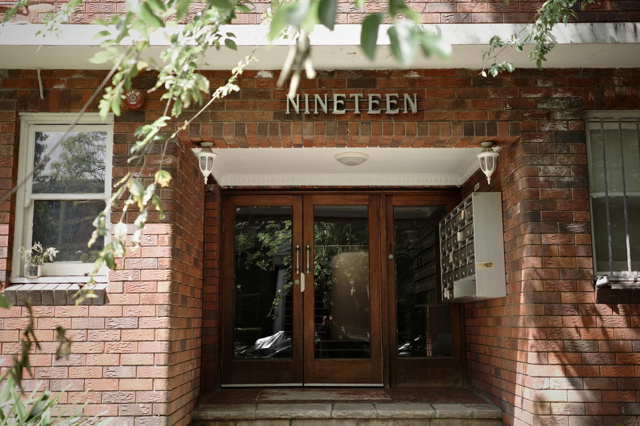 The entrance of a brick building with the word NINETEEN above the door, flanked by windows and surrounded by greenery