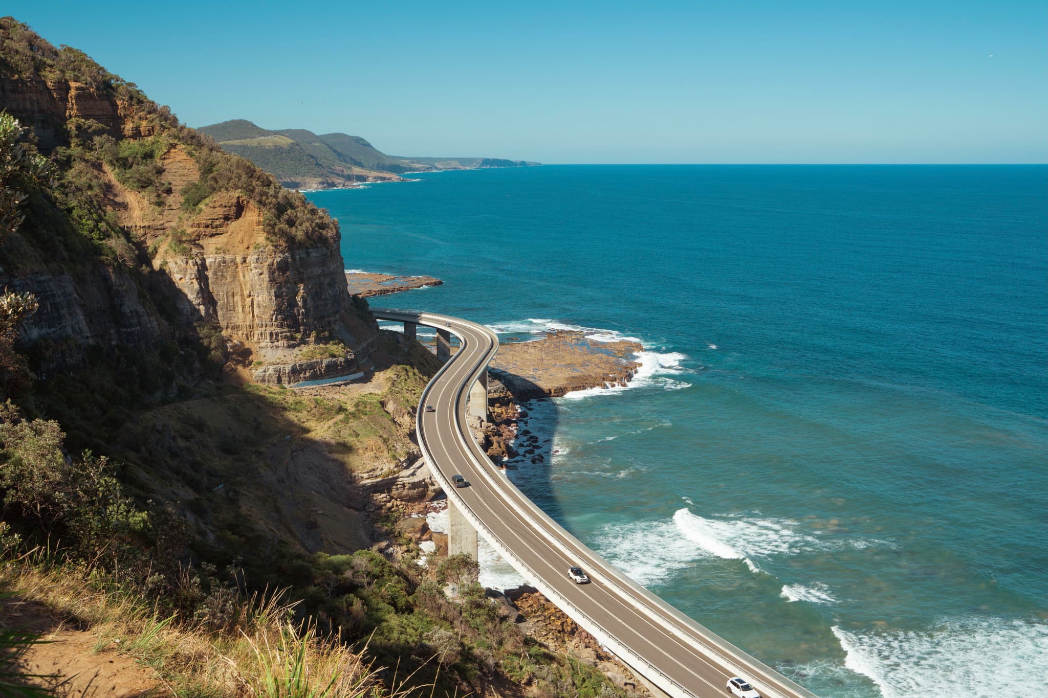 A coastal road curves along the edge of a cliff, with the ocean on one side and rugged terrain on the other, under a clear blue sky