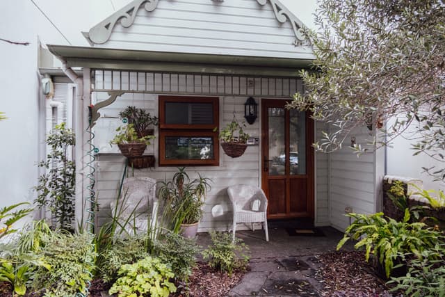 A quaint, small house with a wooden door, decorative trim, and potted plants on the porch, surrounded by greenery