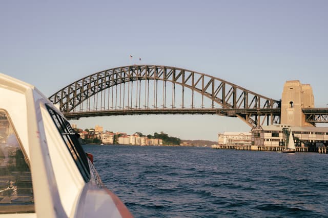 A view of a large steel arch bridge over a body of water, with a boat in the foreground and buildings in the background