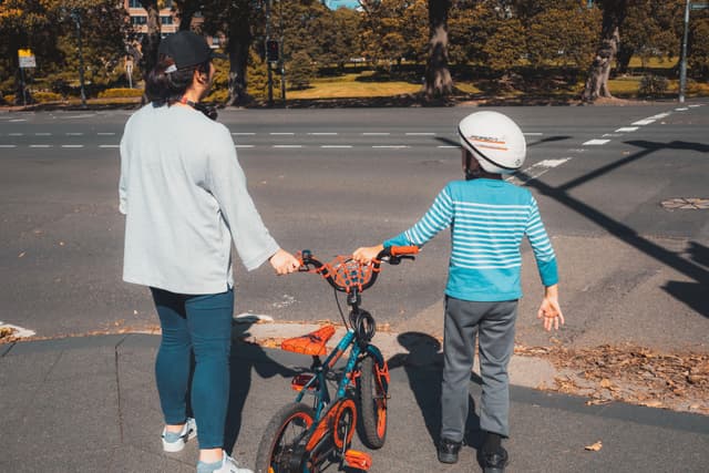 A woman and a child stand at a crosswalk, holding a bicycle. The child wears a helmet, and they are both looking across the street