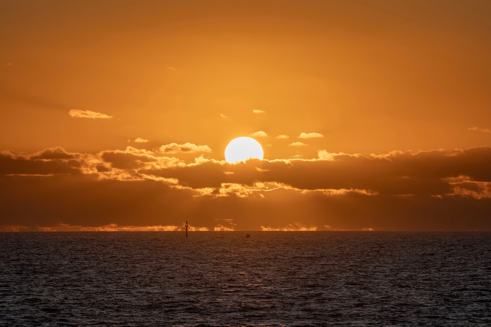 A sunset over the ocean with the sun partially obscured by clouds, casting an orange glow across the sky and water