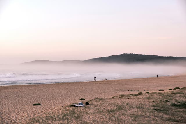 A serene beach scene with a sandy shore, gentle waves, and a misty horizon. A few people are walking along the beach, and hills are visible in the background under a soft, pastel sky