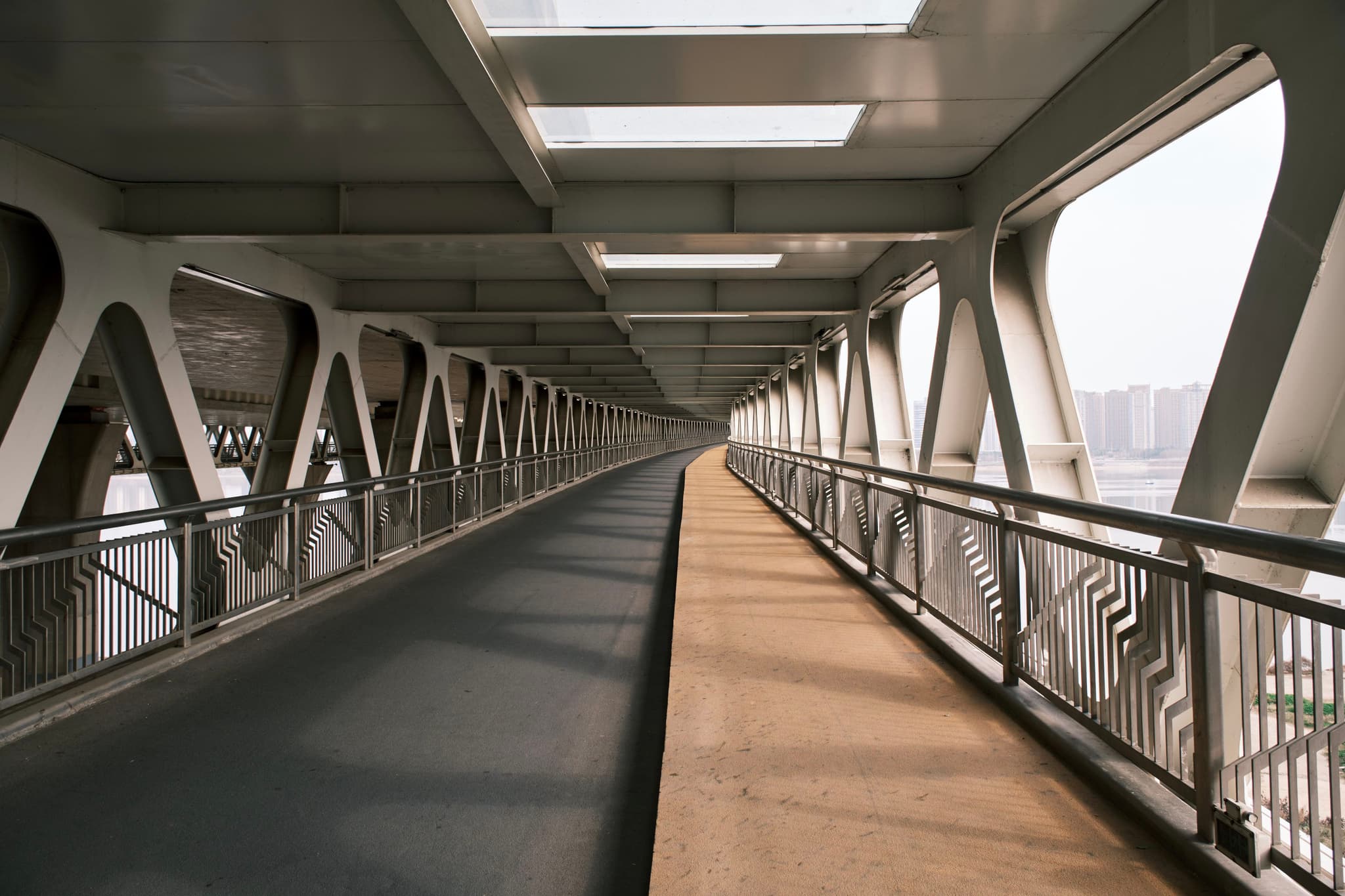A covered pedestrian walkway with metal railings and large windows, featuring a divided path for walking and cycling