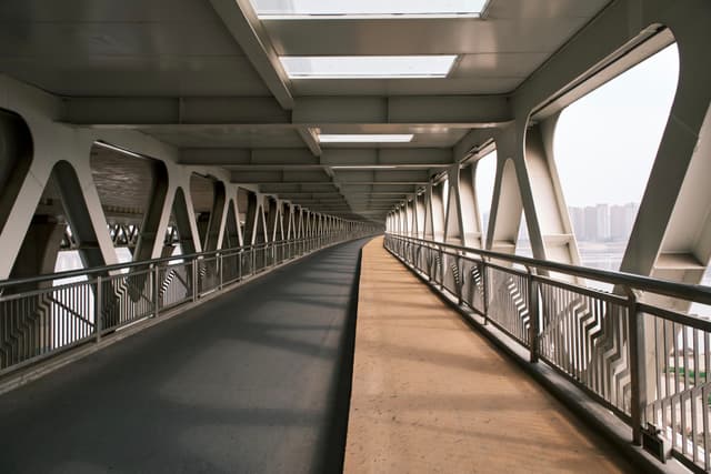 A covered pedestrian walkway with metal railings and large windows, featuring a divided path for walking and cycling