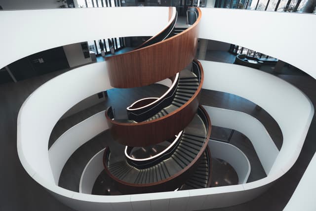 A modern, spiral staircase with wooden steps and a sleek design, surrounded by white walls in a multi-story building