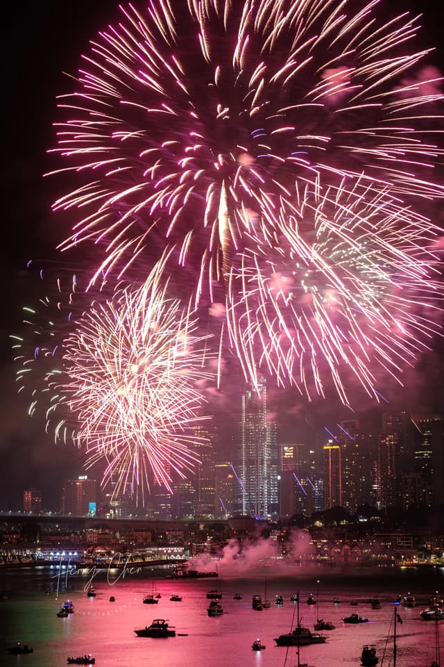 A vibrant fireworks display illuminates the night sky over a cityscape, with reflections on the water and boats visible below