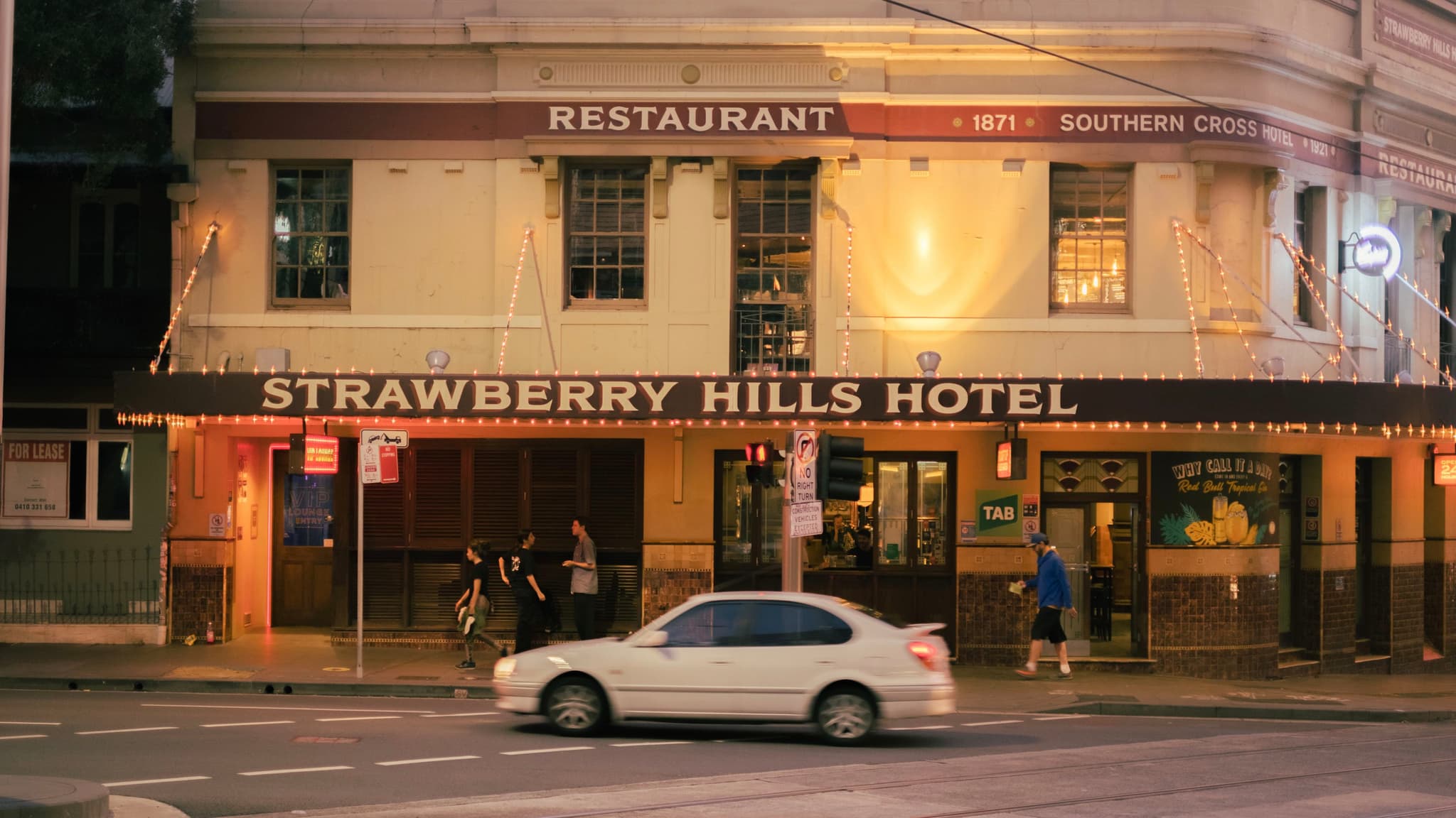 A street view of the Strawberry Hills Hotel with a white car parked in front and a few people walking nearby. The building has a restaurant sign above and is lit warmly