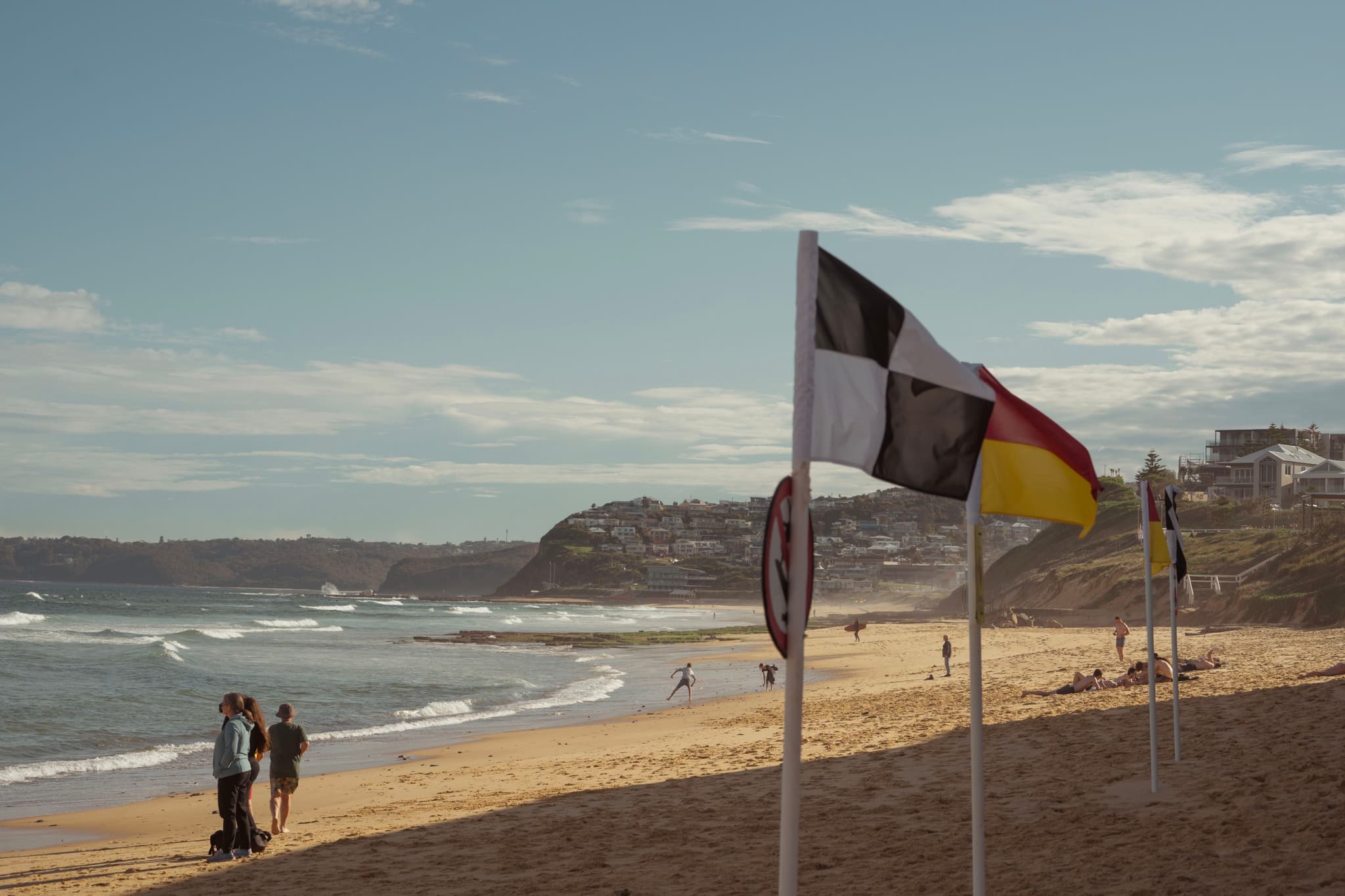 A sandy beach with people walking along the shore, flags marking a designated swimming area, and waves crashing in the background under a partly cloudy sky