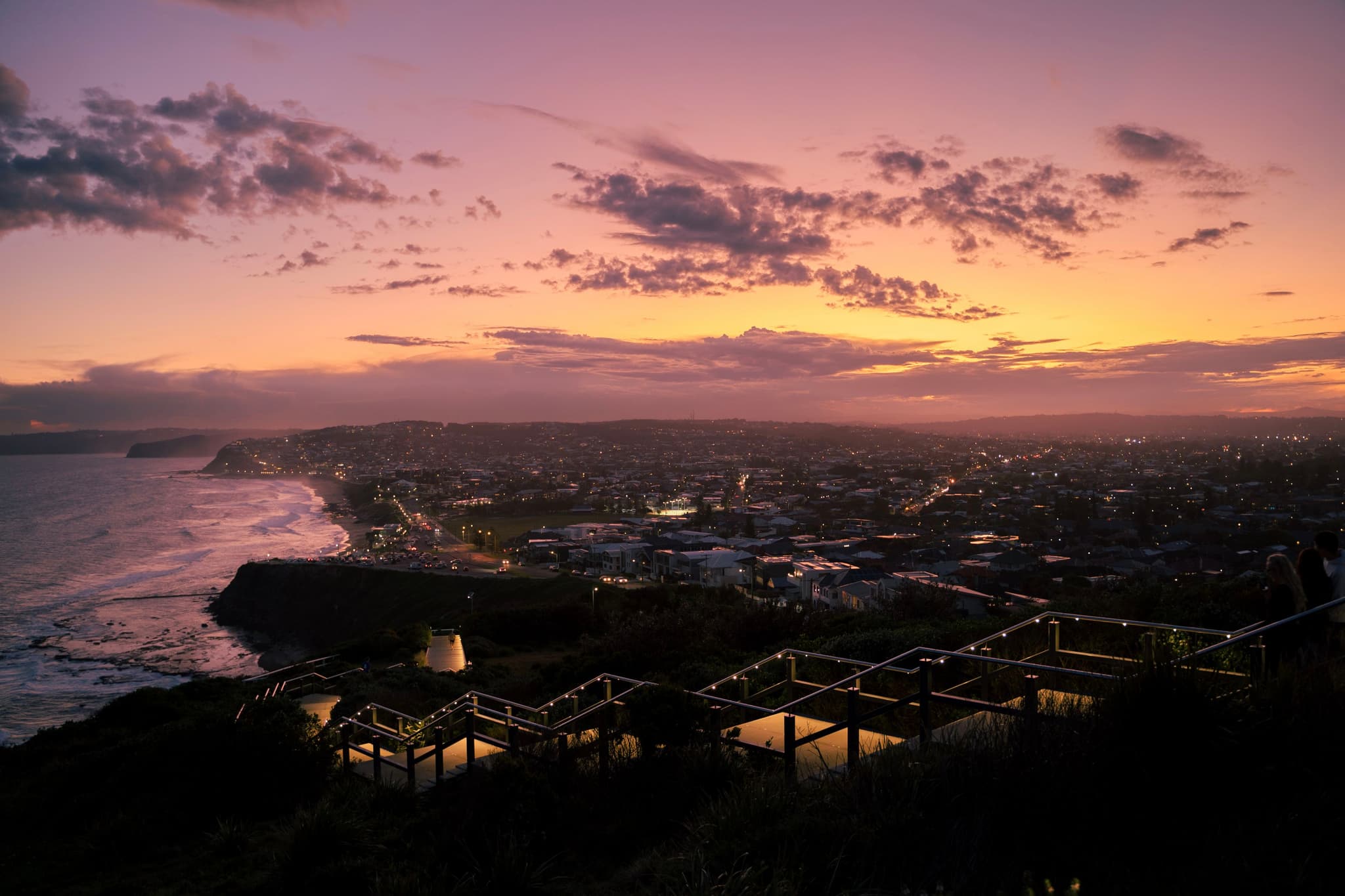 A coastal cityscape at sunset with a vibrant sky, scattered clouds, and a wooden walkway leading down to the shoreline
