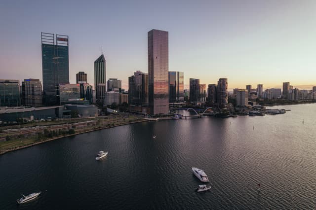 A city skyline at dusk with tall buildings reflecting in the water, and several boats on the river