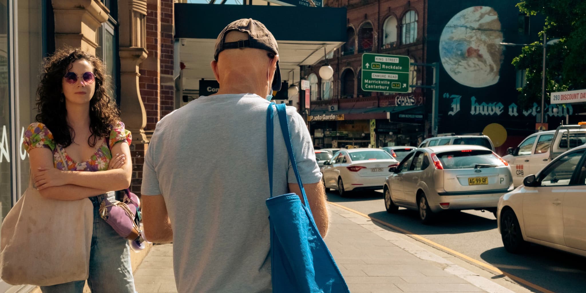 A busy urban street with people walking, cars driving, and a large advertisement on a building in the background