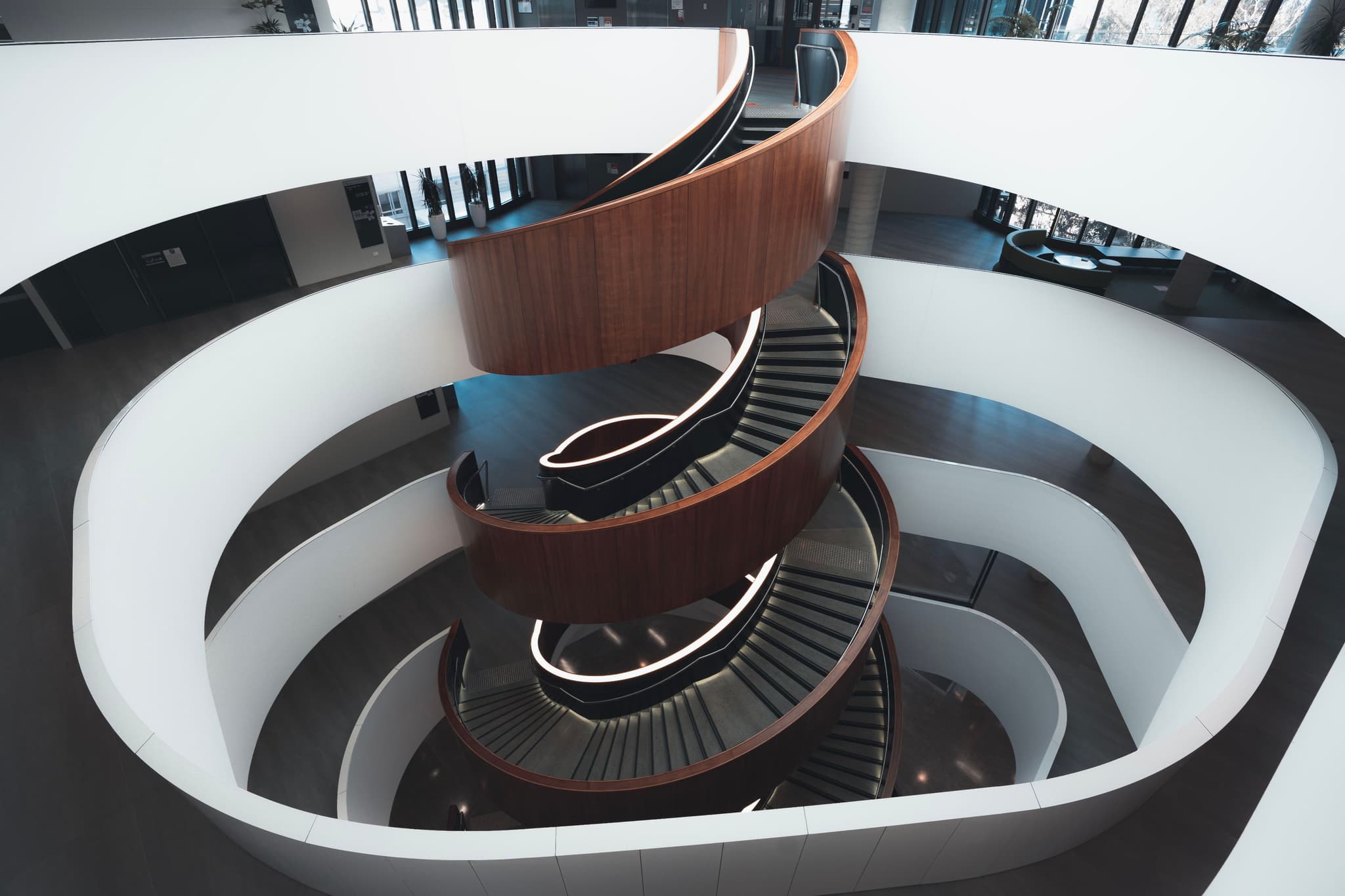 A modern, spiral staircase with wooden steps and a sleek design, surrounded by white walls in a multi-story building