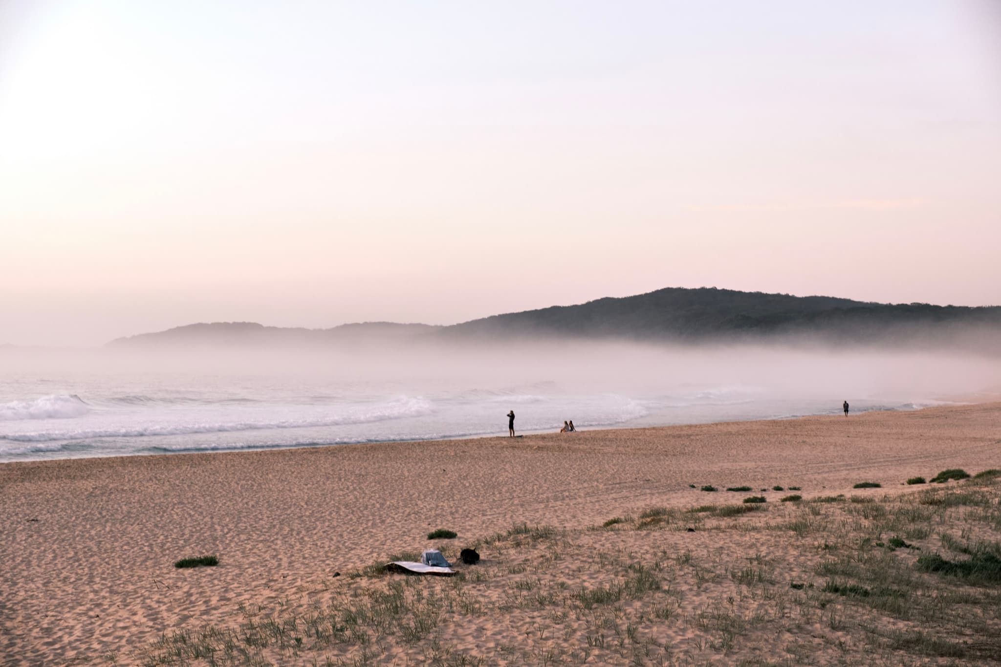 A serene beach scene with a sandy shore, gentle waves, and a misty horizon. A few people are walking along the beach, and hills are visible in the background under a soft, pastel sky