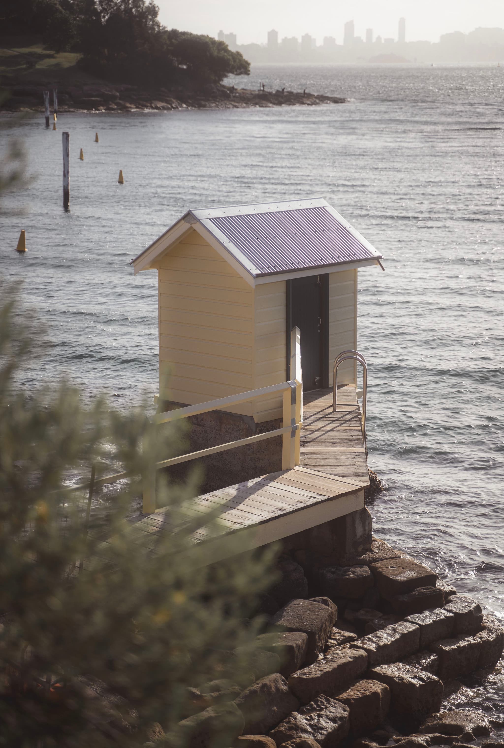 A small wooden shed with a slanted roof sits on a rocky shoreline, connected by a narrow walkway. The water is calm, and a distant city skyline is visible across the bay