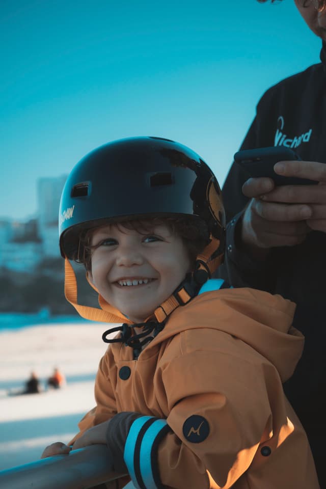 A young child wearing a helmet and an orange jacket smiles while standing next to an adult holding a phone. The background is a snowy landscape with a clear blue sky