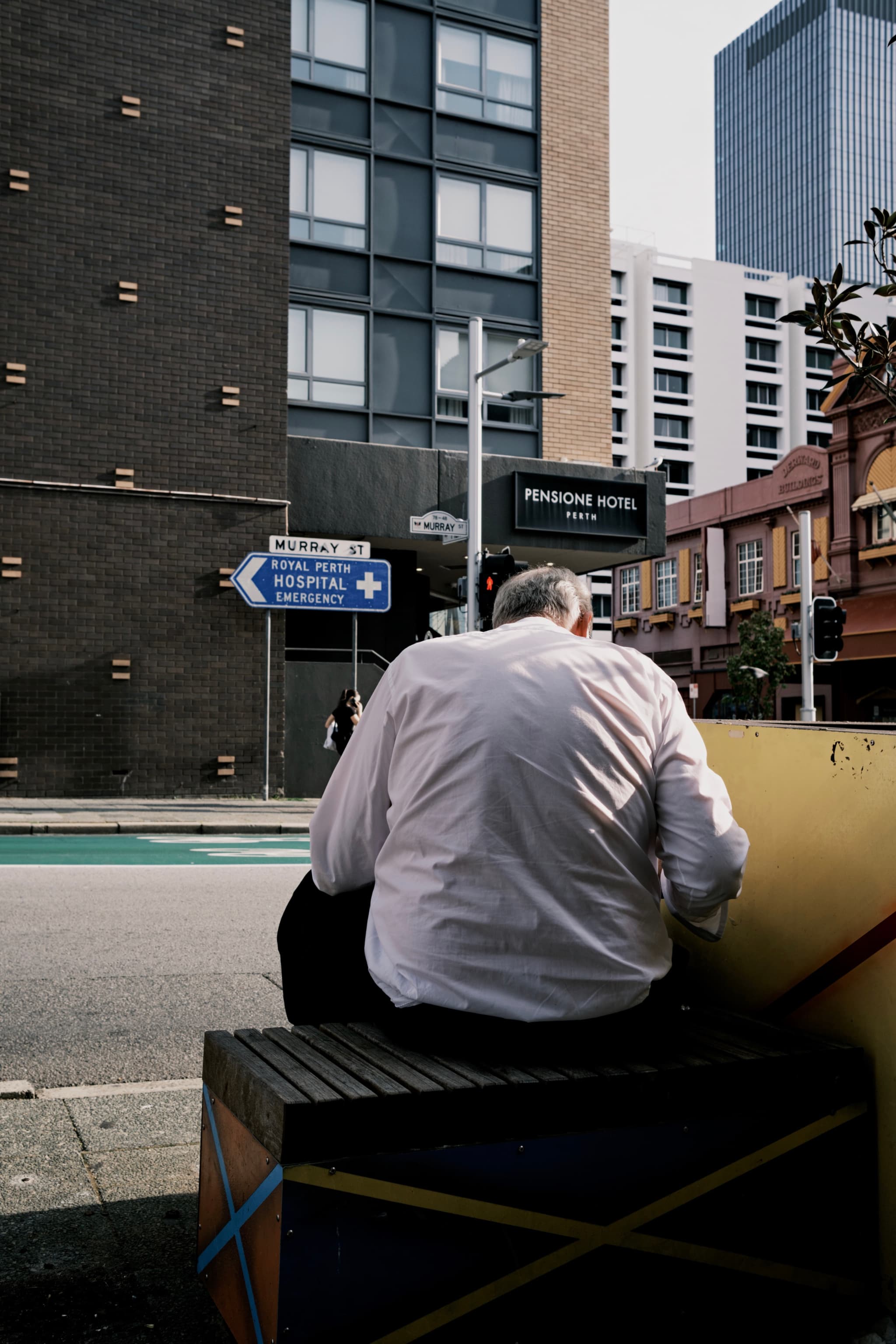 A person in a white shirt sits on a colorful bench near a street intersection, with buildings and a road sign in the background