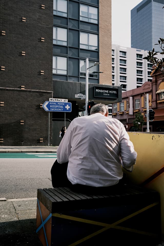 A person in a white shirt sits on a colorful bench near a street intersection, with buildings and a road sign in the background