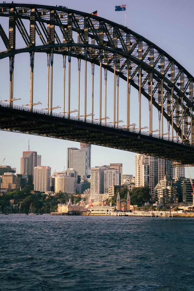 A large steel arch bridge spans across a body of water with a city skyline in the background, featuring modern high-rise buildings