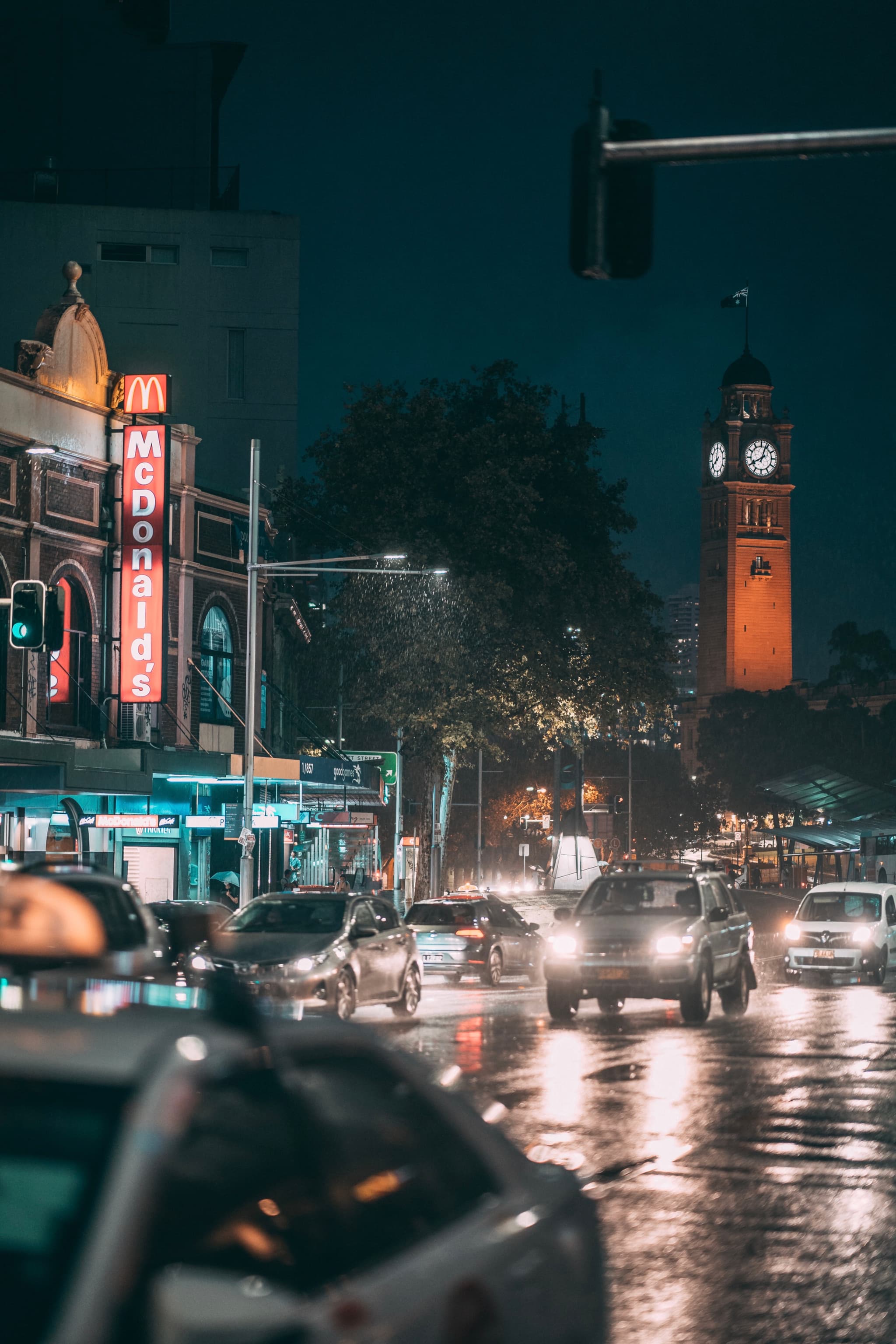 A rainy city street at night with cars, a McDonald's sign, and a clock tower in the background