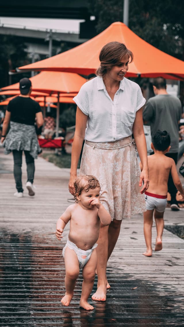 A woman walking with a toddler in a diaper on a wet boardwalk, with orange umbrellas and other people in the background