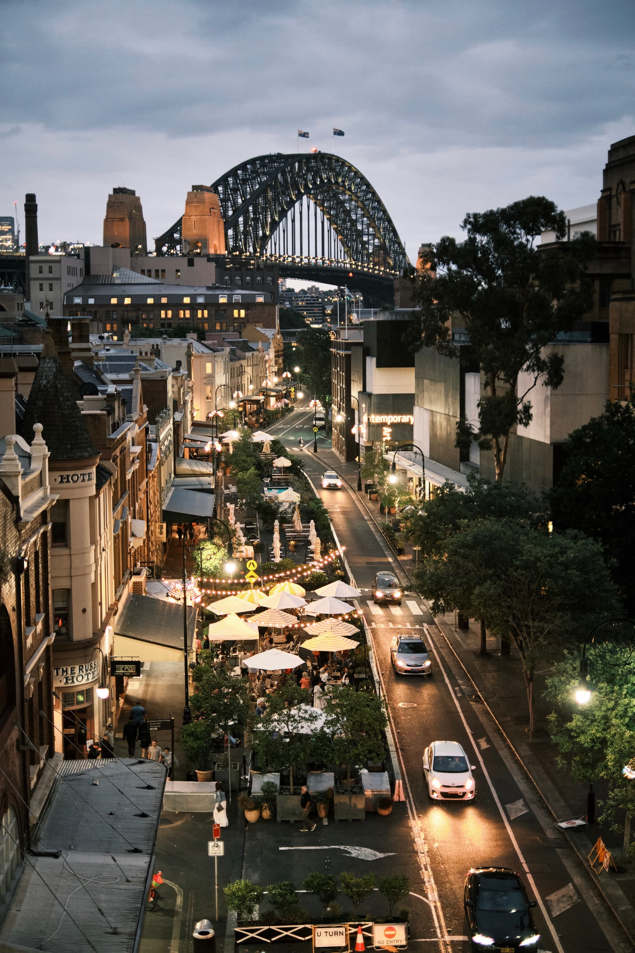 A bustling urban street scene at dusk with cars, pedestrians, and outdoor dining, set against the backdrop of a large, illuminated bridge
