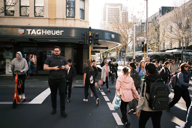 A busy city street with people crossing at a pedestrian crosswalk. A TAG Heuer store is visible in the background, and a person is riding an electric scooter