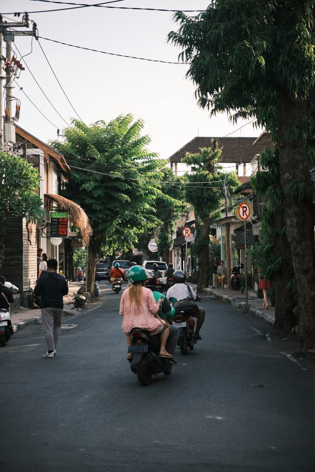 A street scene with people riding scooters and a pedestrian walking, surrounded by trees and buildings