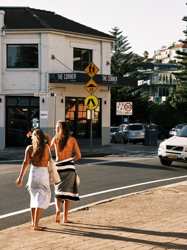 Two people in beach attire walk along a street near a corner building with signs and parked cars