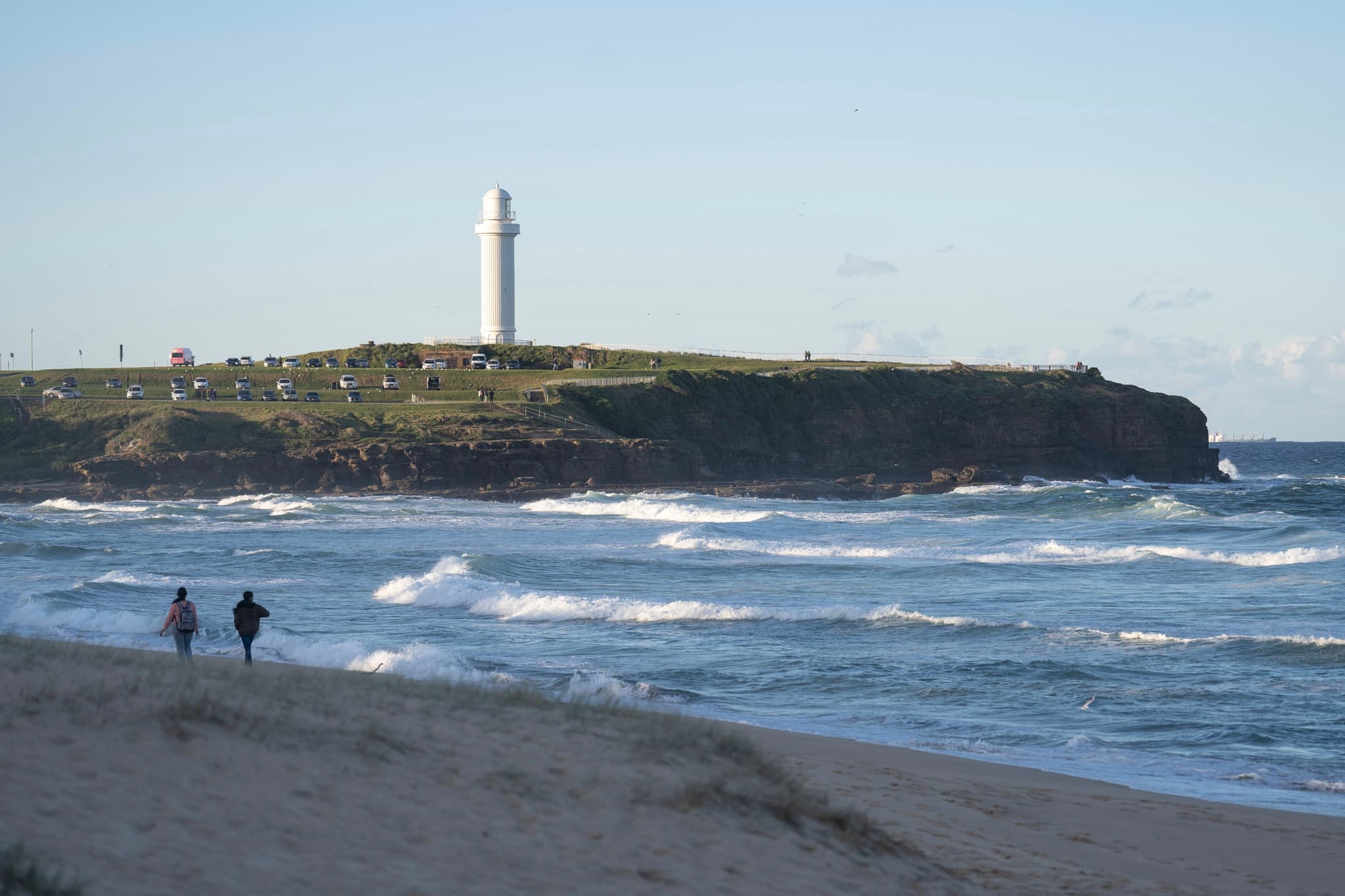 A lighthouse stands on a grassy cliff overlooking a beach with two people walking along the shoreline and waves crashing onto the sand