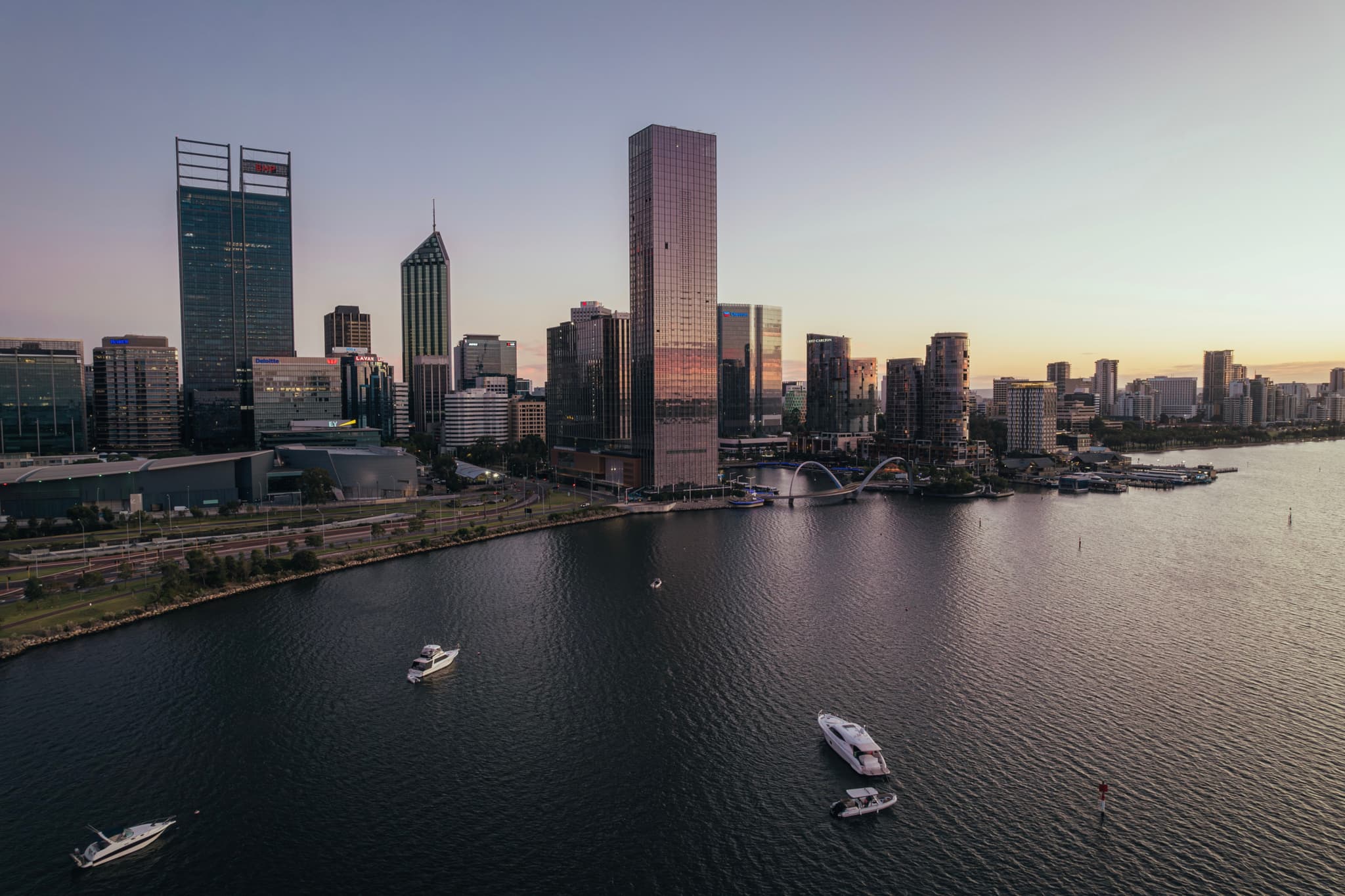 A city skyline at dusk with tall buildings reflecting in the water, and several boats on the river