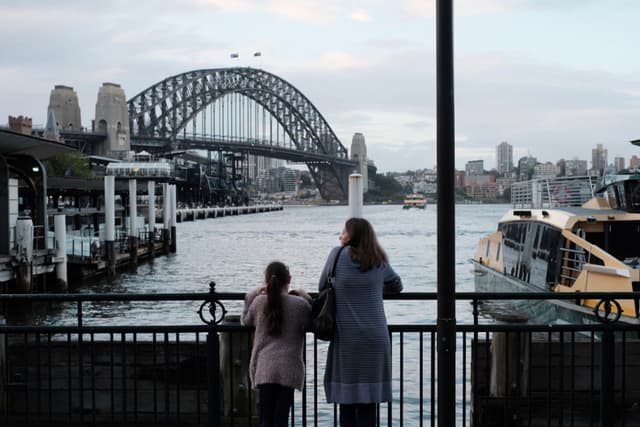 Two people stand by a railing overlooking a body of water with the Sydney Harbour Bridge in the background. A ferry is visible on the right, and the city skyline is in the distance