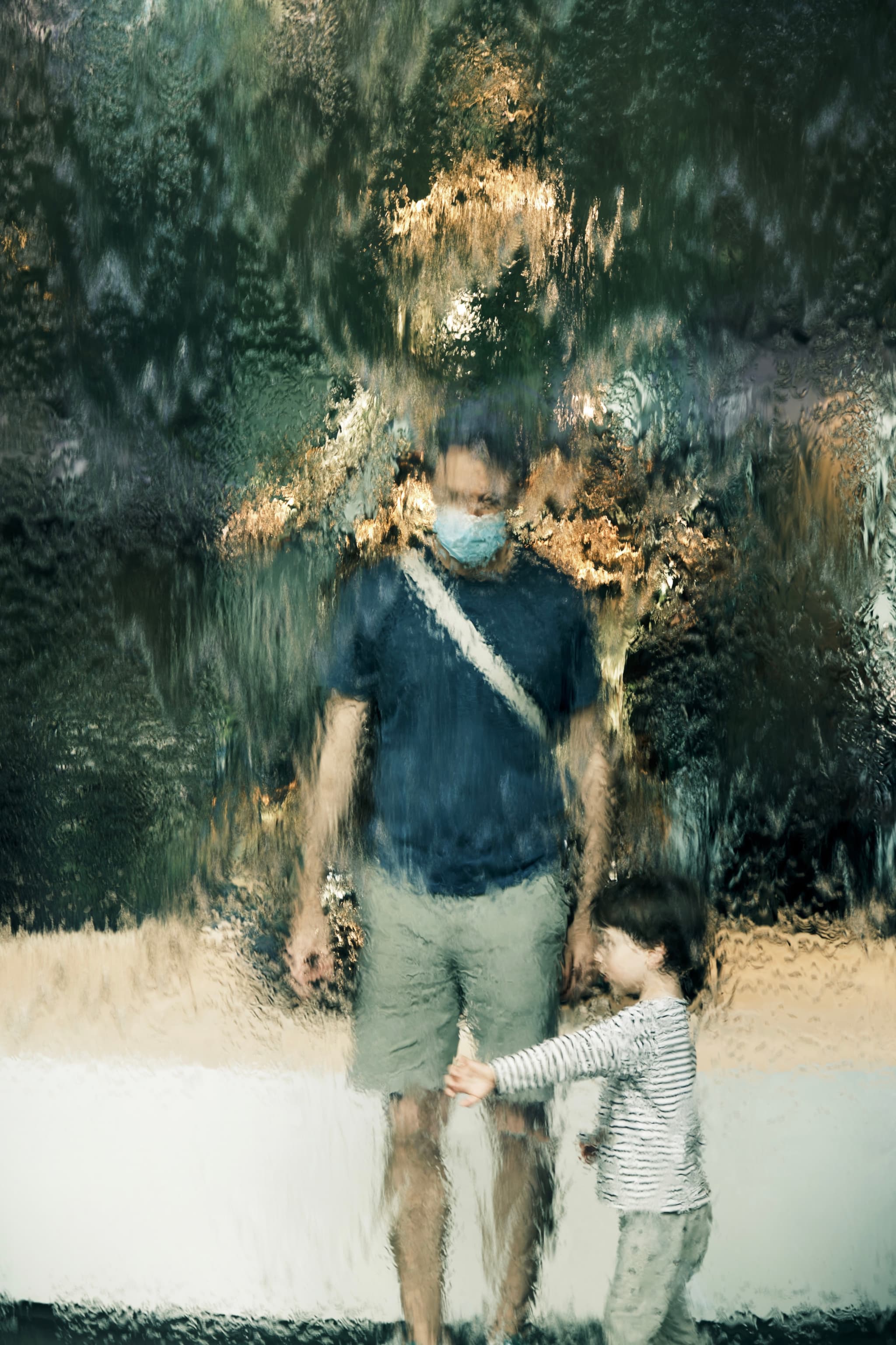 A man wearing a mask and a child stand behind a glass wall with water streaming down, creating a blurred effect