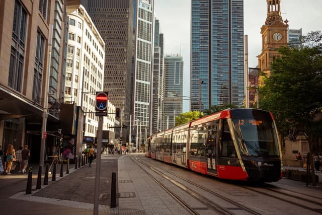 A modern tram travels through a city street flanked by tall buildings, with pedestrians walking along the sidewalks