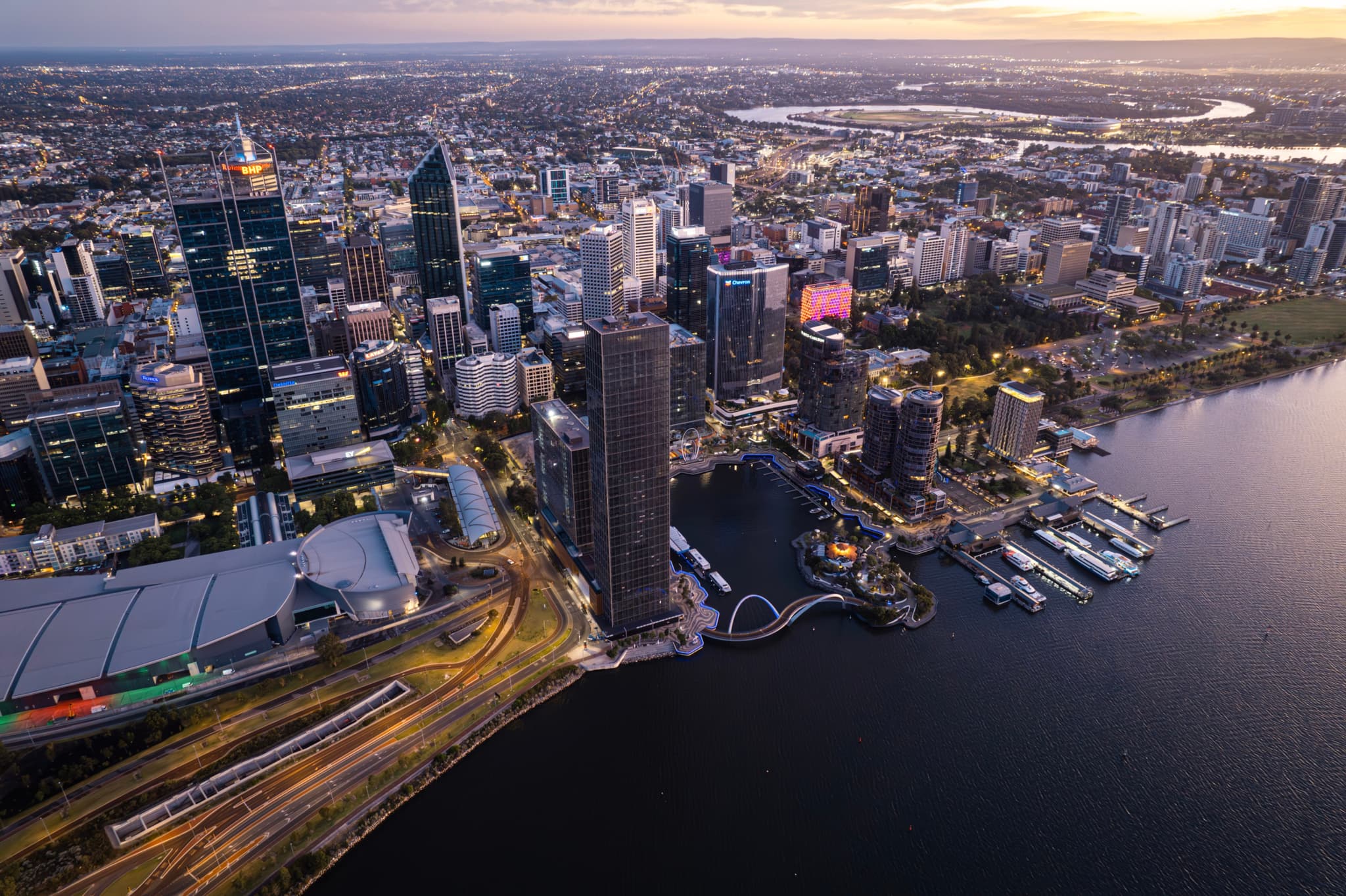 Aerial view of a cityscape with tall buildings, a waterfront, and a network of roads, captured during sunset