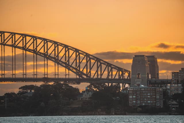 A bridge silhouetted against an orange sunset sky, with buildings and trees in the foreground