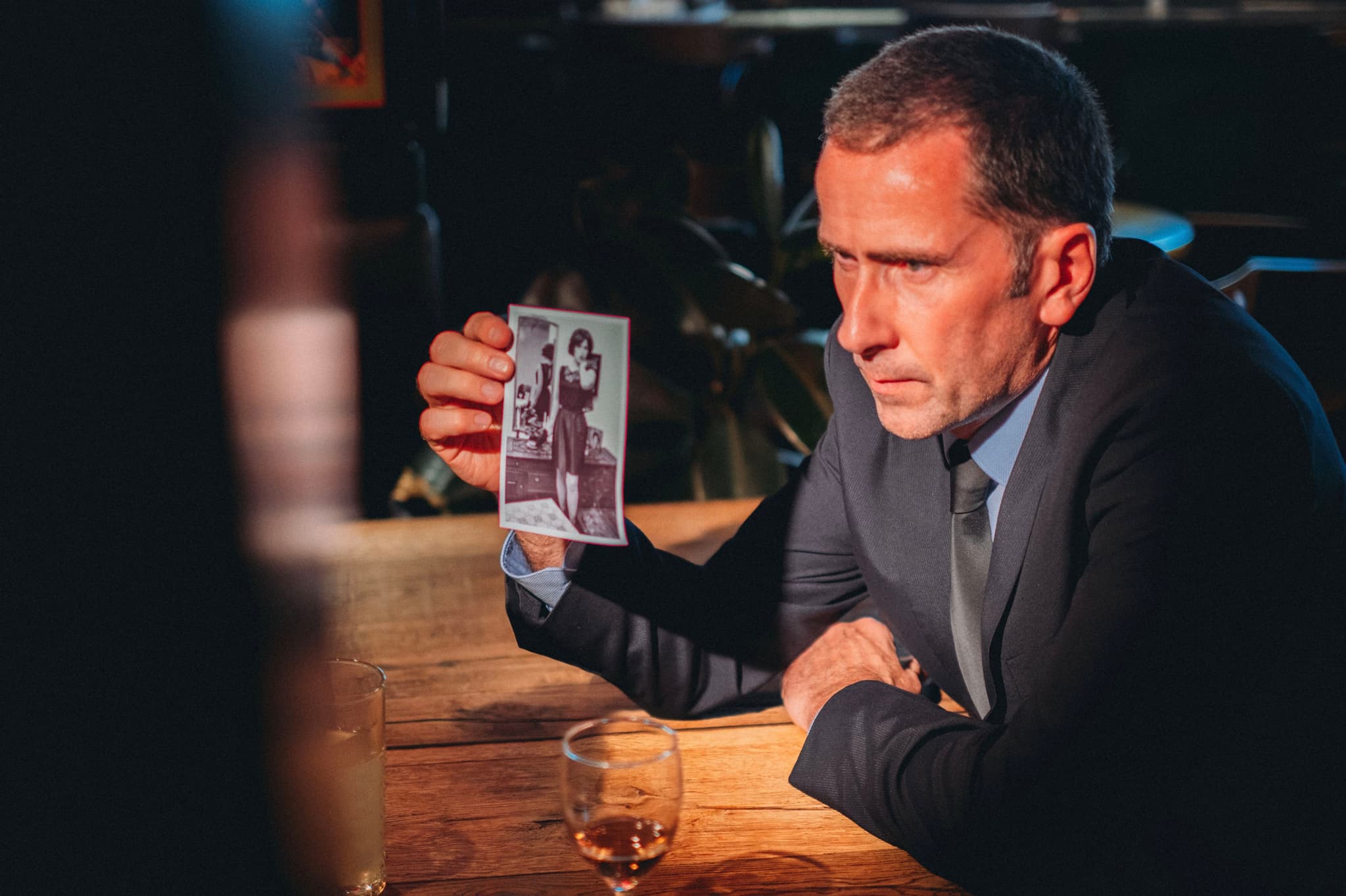 A man in a suit sits at a wooden table, holding a photograph and looking intently at it, with a glass of whiskey nearby