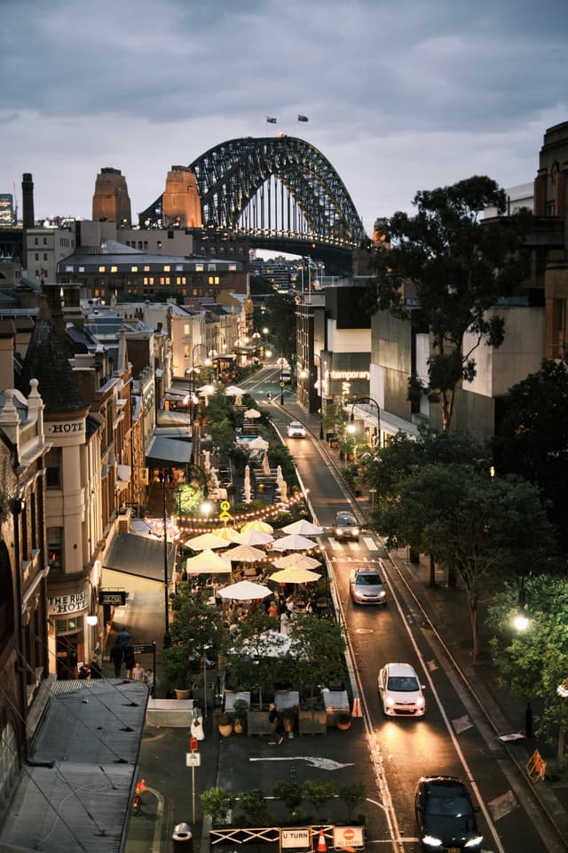 A bustling urban street scene at dusk with cars, pedestrians, and outdoor dining, set against the backdrop of a large, illuminated bridge
