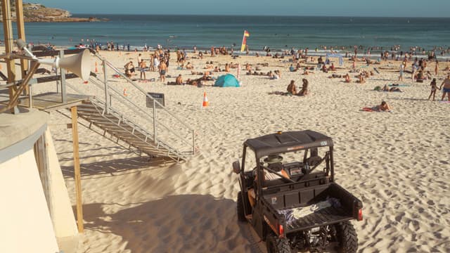 A crowded beach with people sunbathing and swimming, a lifeguard vehicle parked on the sand, and a clear blue sky